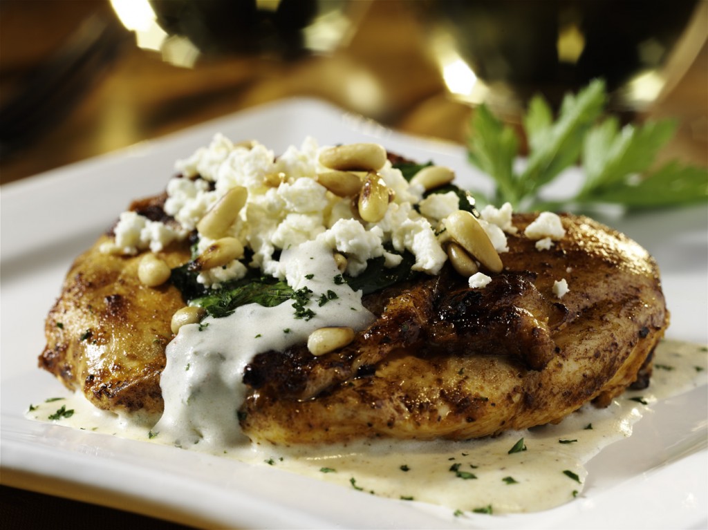 Broiled chicken with pine nuts and creamy white sauce sits on a white plate. Close-up shot.