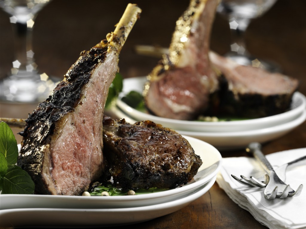 Close up of lamb chops on white plates. Silverware sits on white paper napkins in the foreground. There are crystal wine glasses blurred out in the background.